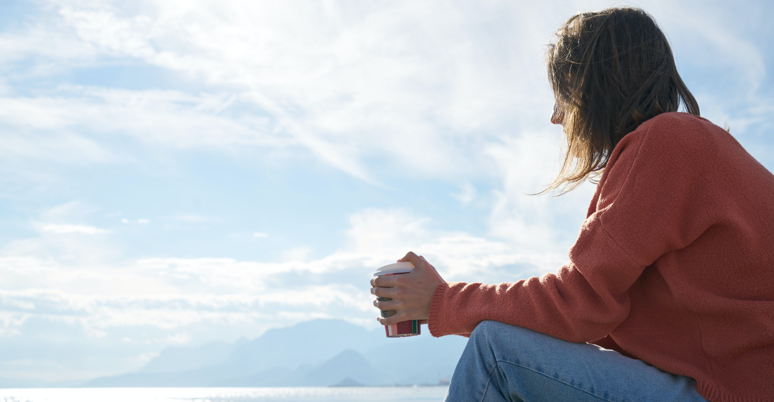 Eine Frau sitzt mit einem Kaffee in der Hand nachdenklich am Strand.