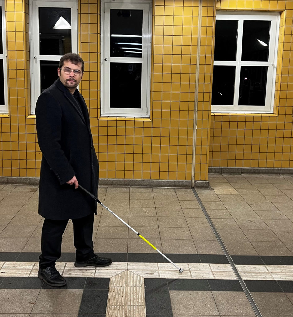 Der TikToker Erdin aka Mr. BlindLife mit seinem Blindenstock in einer U-Bahn-Station, die gelb gefliest ist. Er folgt mit seinem Stock dem Bodenleitsystem und schaut dabei in die Kamera.