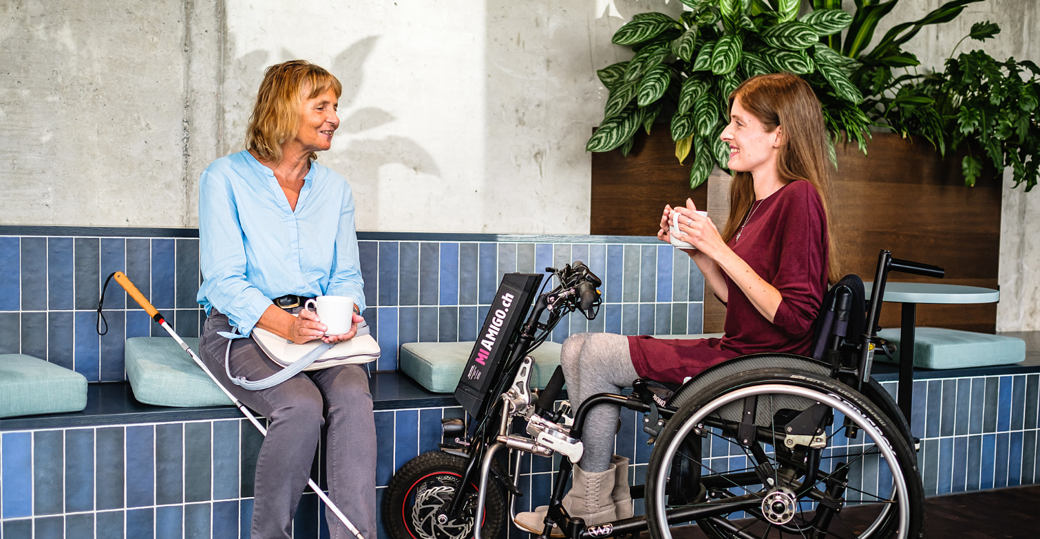 Zwei Frauen, eine mit Blindenstock und eine Frau im Rollstuhl, unterhalten sich in einem gefliesten Raum miteinander.