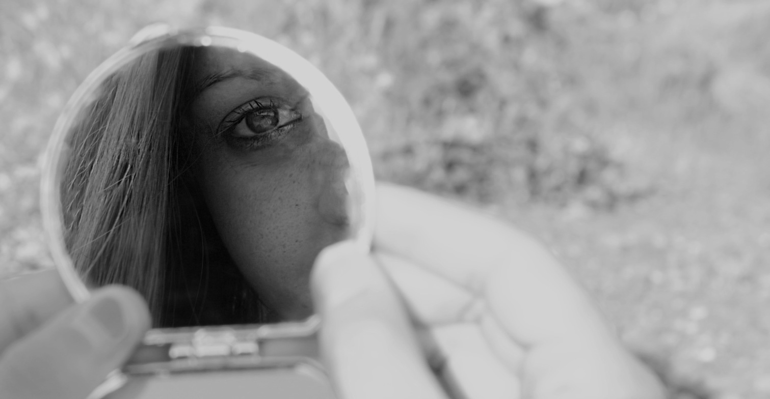 Eine Frau sieht in einen runden Spiegel, ihr Auge und ein Teil ihrer Haare sind sichtbar.