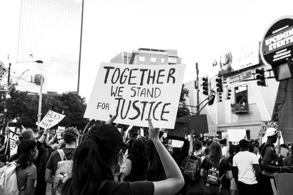 Eine schwarzweiß Fotografie von einer Black Lives Matter-Demonstration zeigt Demonstrierende von hinten mit verschiedenen Schildern. Im Vordergrund hält eine Person ein Schild mit dem Slogan: "Together we stand for Justice".