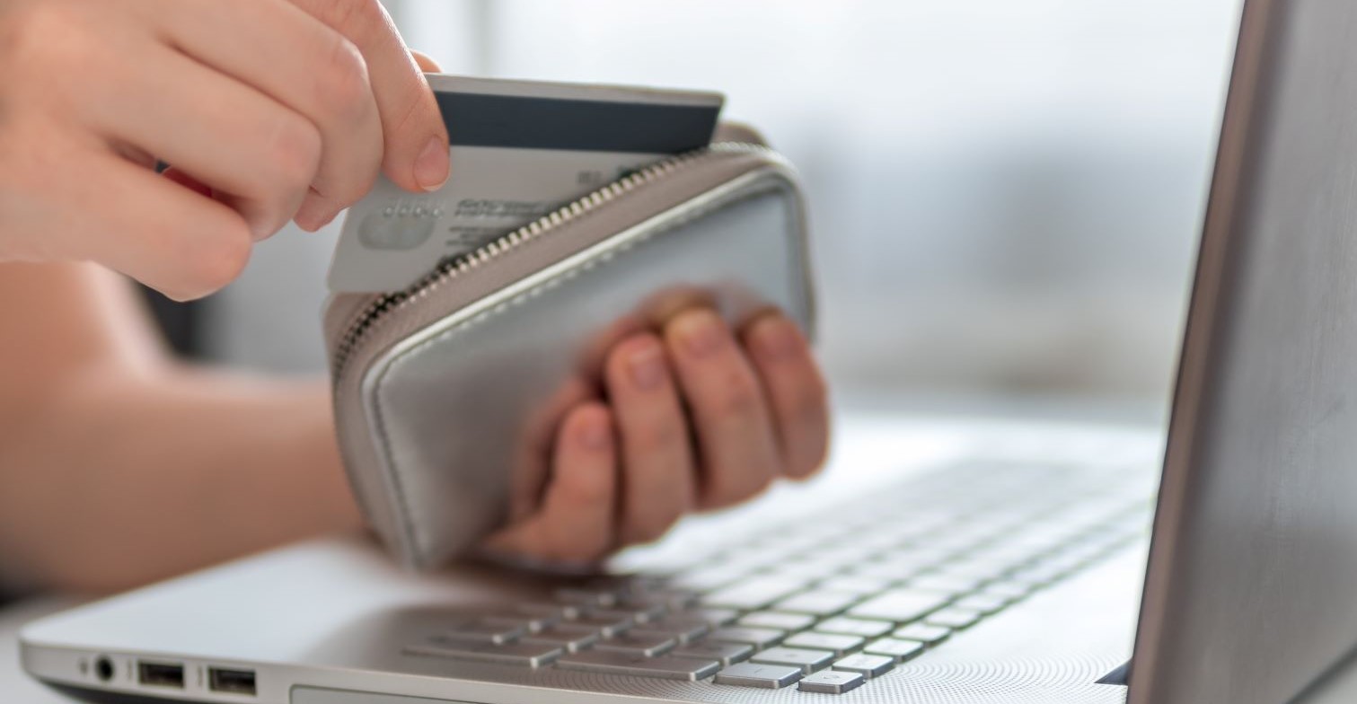 Auf dem Foto ist ein Computer zu sehen. Ein Person sitzt davor und nimmt eine Bankkarte aus dem Portemonnaie.