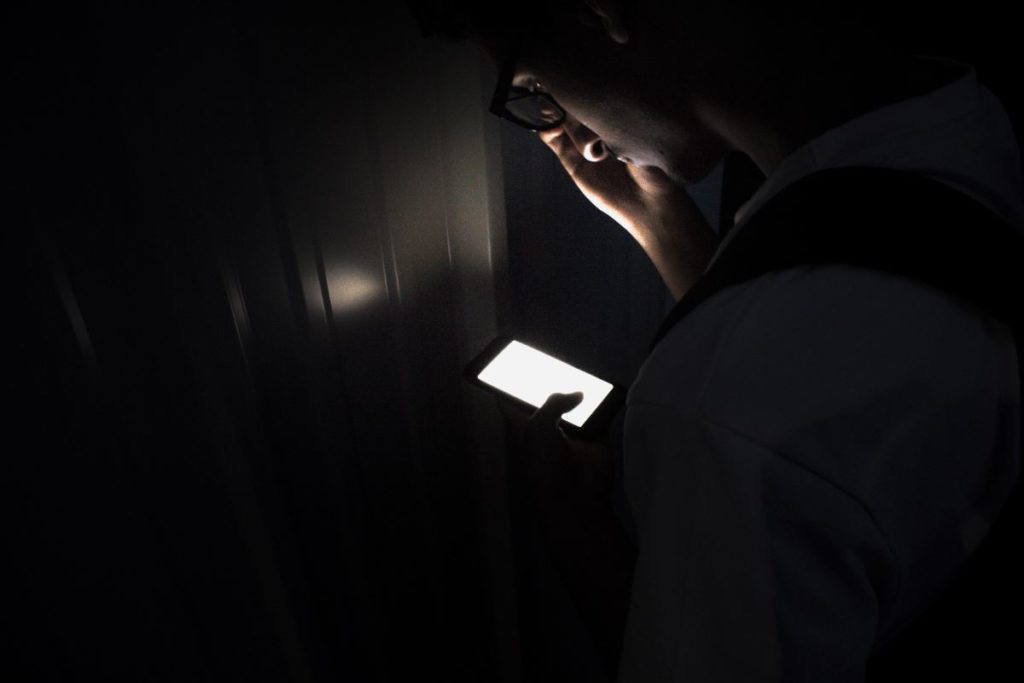Person steht im Dunkeln vor einer Zimmertür mit einem Handy in der Hand.