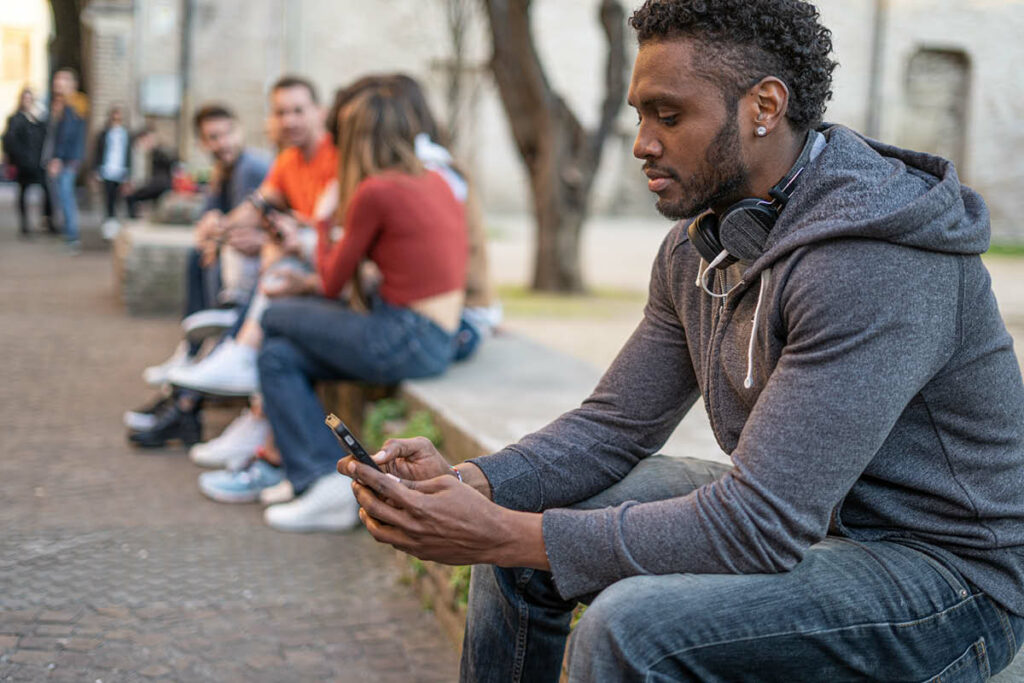 Rassismus im Internet: Männliche BIPoC Person sitzt neben einer Gruppe und schaut in ein Smartphone