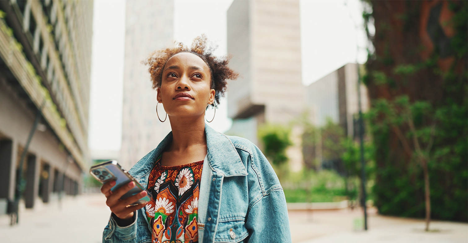 Bild einer jungen Schwarzen Frau in Jeansjacke mit Handy in der Hand vor einer Hochhaus-Kulisse