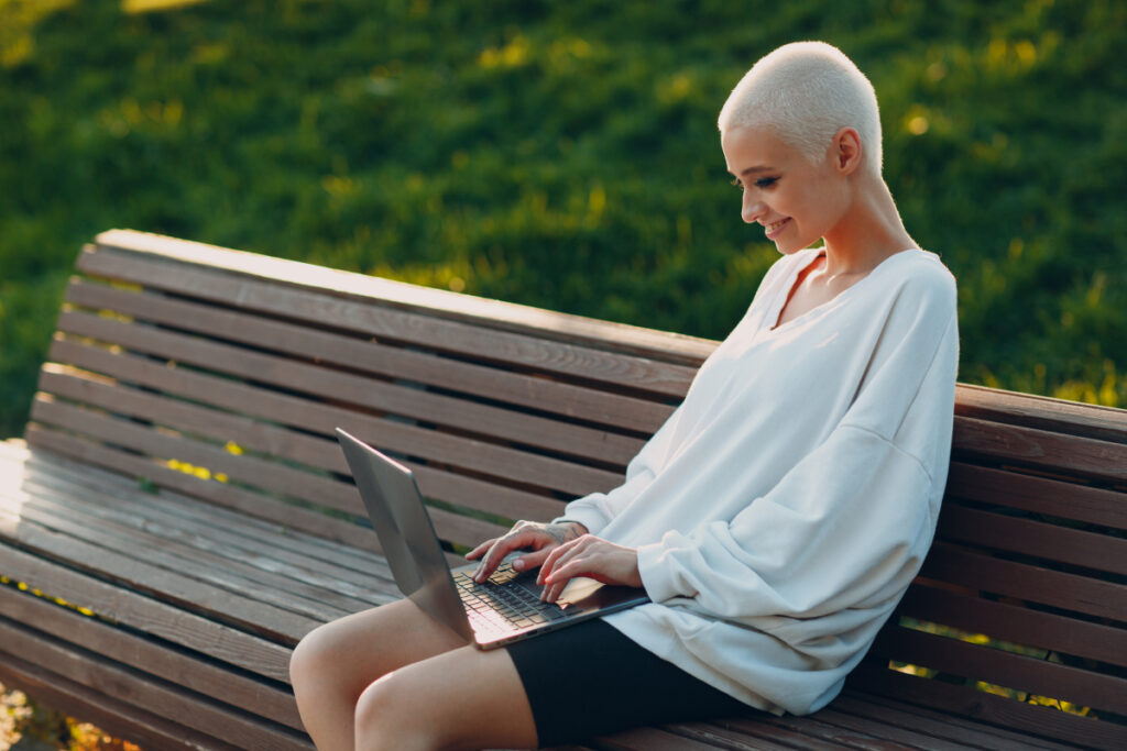 Macht der Onlinekommentare - Lächelnde Frau sitzt auf Parkbank und tippt auf der Laptoptastatur
