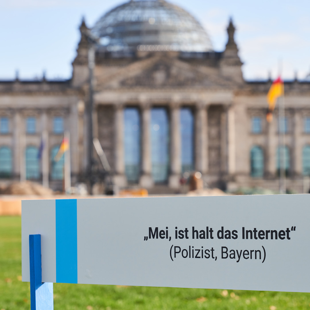 HateAid Hürdenlauf zum Gesetz gegen digitale Gewalt - Aktion vor dem Bundestag. Zu sehen ist im Hintergrund das Reichstagsgebäude sowie im Vordergrund ein Schild mit der Aufschrift "Mei, ist halt das Internet (Polizist, Bayern)"