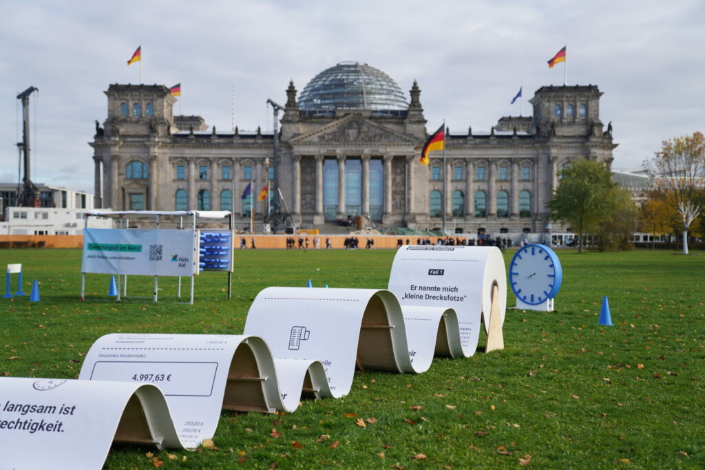 Hürdenlauf mit großer Rechnug, Uhr und einem Gerüst mit Banner zur Petition vor dem Bundestagsgebäude in Berlin.
