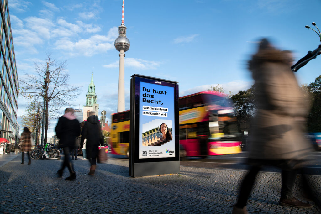 HateAid Menschenrechtskampagne #UnserInternet. Zu sehen ist ein Foto eines belebten Bürgersteigs und ein Werbebildschirm der Firma Wall GmbH. Copyright: Wall GmbH