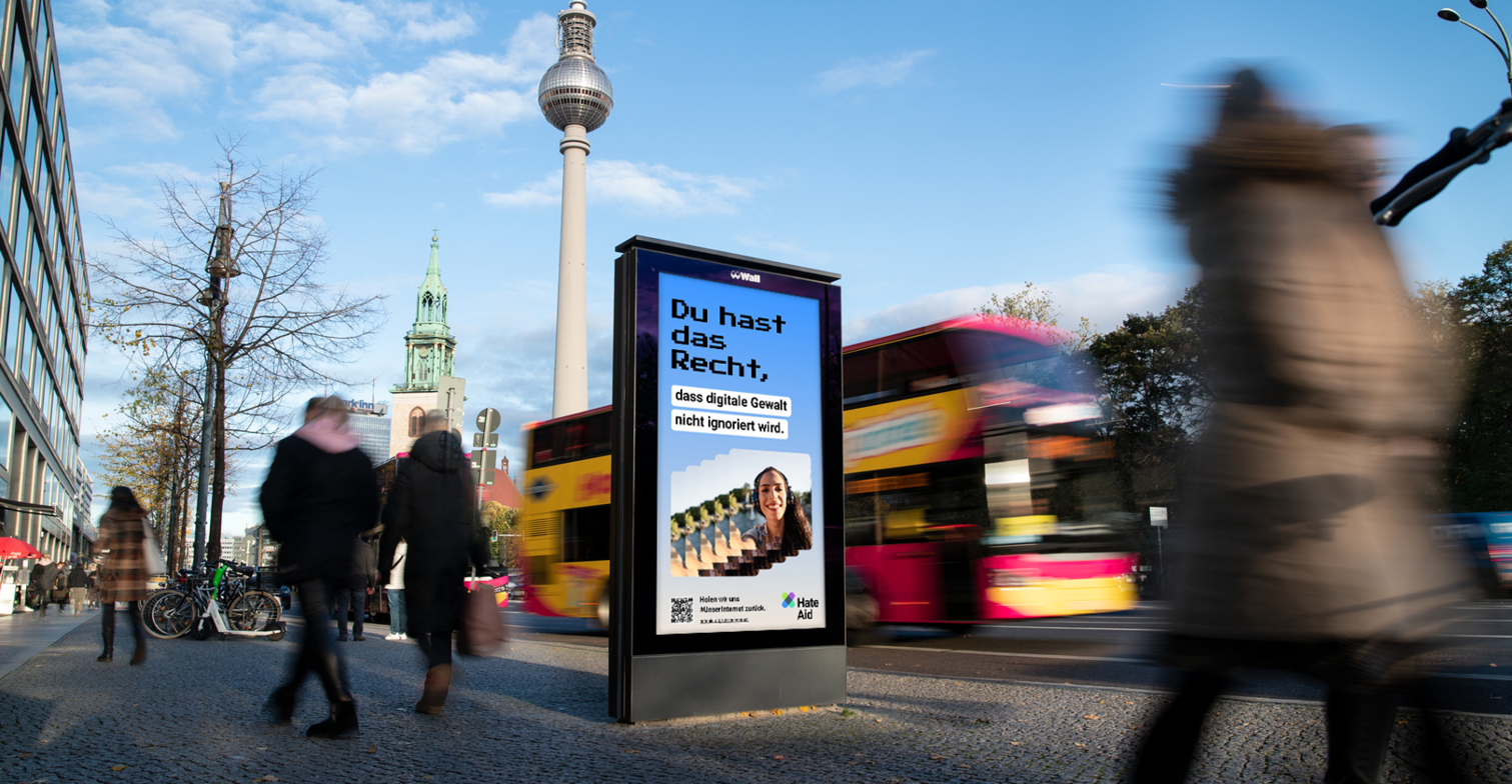 HateAid Menschenrechtskampagne #UnserInternet. Zu sehen ist ein Foto eines belebten Bürgersteigs und ein Werbebildschirm der Firma Wall GmbH. Copyright: Wall GmbH