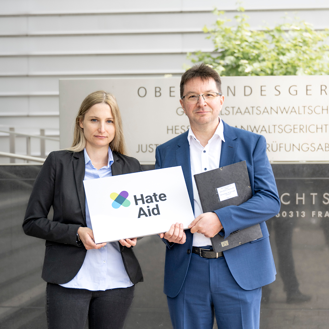 Josephine Ballon und Dr. Michael Blume vor dem Oberlandesgericht Frankfurt am Main. Beide halten gemeinsam ein weißes Schild mit der Aufschrift "HateAid" in den Händen.