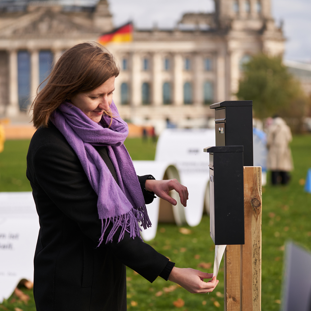 Zu sehen ist eine Frau vor dem Reichstagsgebäude, die ein Schreiben in einen Briefkasten mit nicht vorhandenem Boden wirft und unten wieder auffängt.