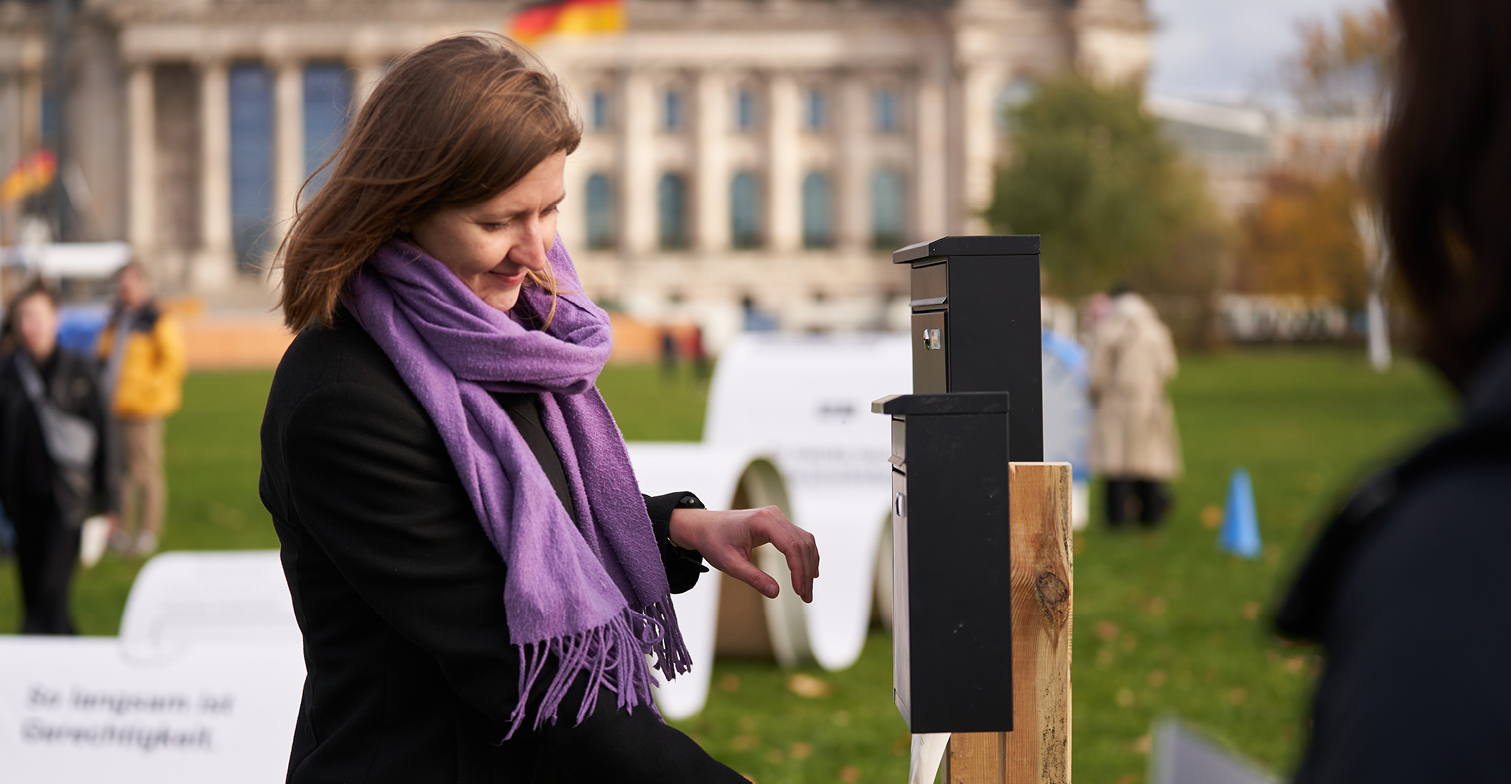 Zu sehen ist eine Frau vor dem Reichstagsgebäude, die ein Schreiben in einen Briefkasten mit nicht vorhandenem Boden wirft und unten wieder auffängt.